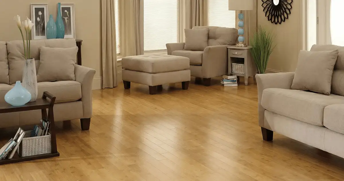 A living room with professionally installed bamboo flooring, beige sofas, a matching ottoman, a side table with a blue lamp, vases, framed pictures, and large windows with blinds and curtains in Saint Petersburg, FL